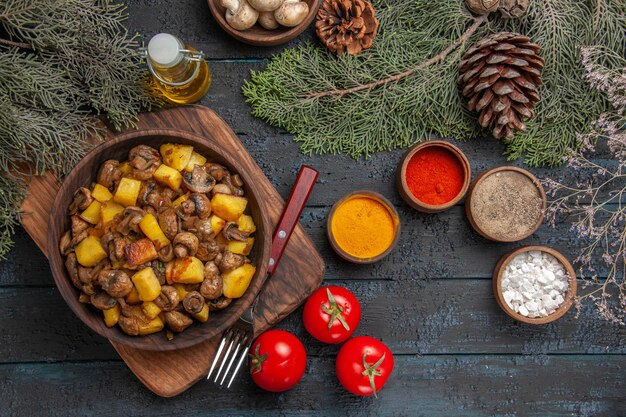 Top view dish and spices dish of potatoes and mushrooms on the cutting board next to the fork three tomatoes and colorful spices under oil bowl of white mushrooms and spruce branches