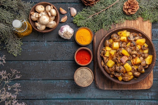Вид сверху блюдо и тарелка со специями из грибов и картофеля на разделочной доске рядом с разноцветным маслом специй в бутылочном чесночном миске с грибами под ветвями с шишками