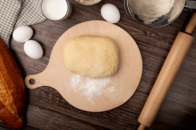 Top view of dough and flour on cutting board with eggs milk rolling pin on wooden background