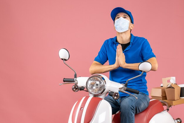 Top view of dreamy courier guy in medical mask wearing hat sitting on scooter delivering orders on pastel peach background