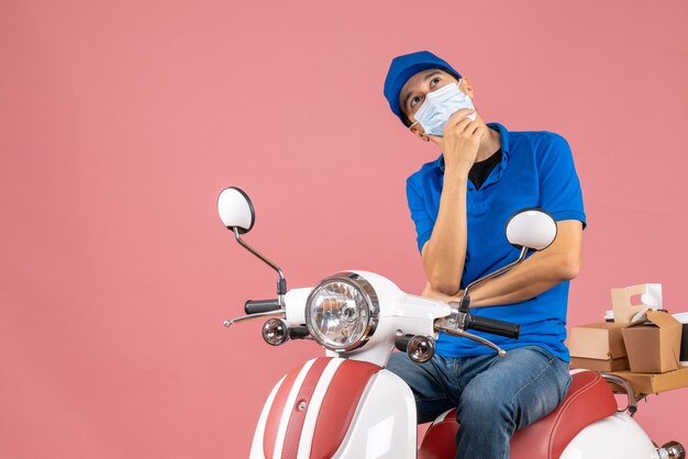 Top view of dreamy delivery guy in medical mask wearing hat sitting on scooter on pastel peach background