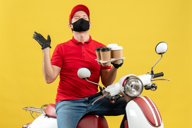 Top view of dreamy delivery man wearing uniform and hat gloves in medical mask sitting on scooter showing orders