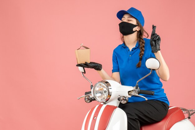 Вид сверху мечтательного доставщика в медицинской маске и перчатках, сидящего на скутере, доставляющего заказы на пастельно-персиковом фоне