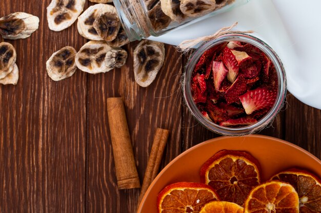 Top view of dried banana chips scattered from a glass jar and dried strawberry slices in a glass jar with dried orange slices on a plate on wooden background