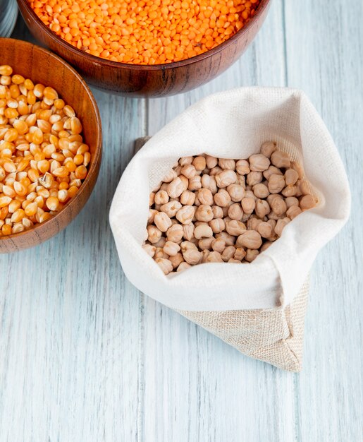 Top view of dried chickpeas in a sackcloth and corn seeds with red lentils in wooden bowls on rustic table