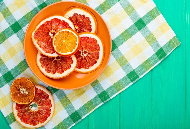 Top view of dried orange and grapefruit slices in a plate on plaid tablecloth on green wooden background with copy space