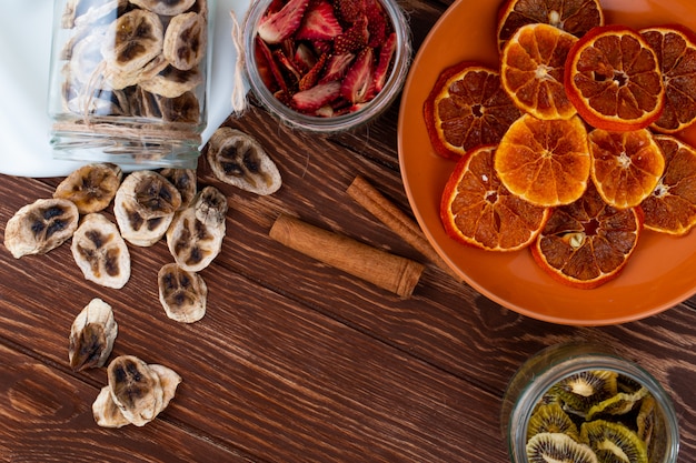 Free photo top view of dried orange slices in a plate and scattered dried banana chips from a glass jar with cinnamon sticks on wooden background