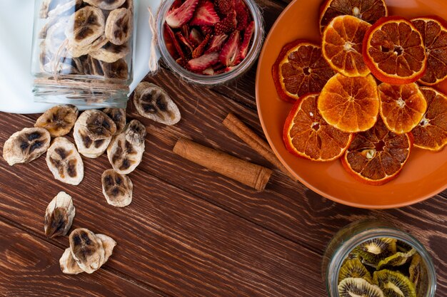 Top view of dried orange slices in a plate and scattered dried banana chips from a glass jar with cinnamon sticks on wooden background