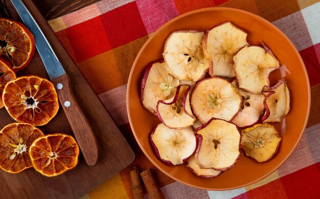 Top view of dried orange slices with kitchen knife on a wooden cutting board and dried apple slices on a plate on plaid tablecloth
