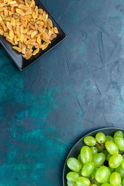 Top view dried raisins inside black form with fresh green grapes on light-blue background.