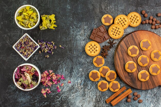 Top view of dry flowers on bowls with cookies on wooden platter and sweets on grey background