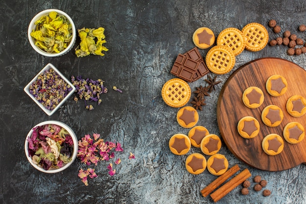 Top view of dry flowers on bowls with cookies on wooden platter and sweets on grey background