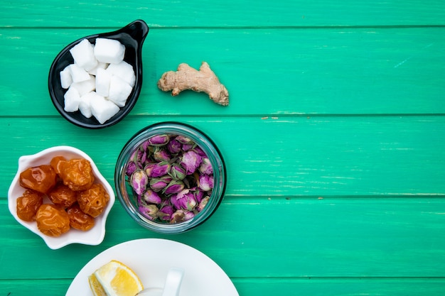 Top view of dry rose buds in a glass jar with dried cherry plums, sugar cubes and a cup of tea on green wood with copy space