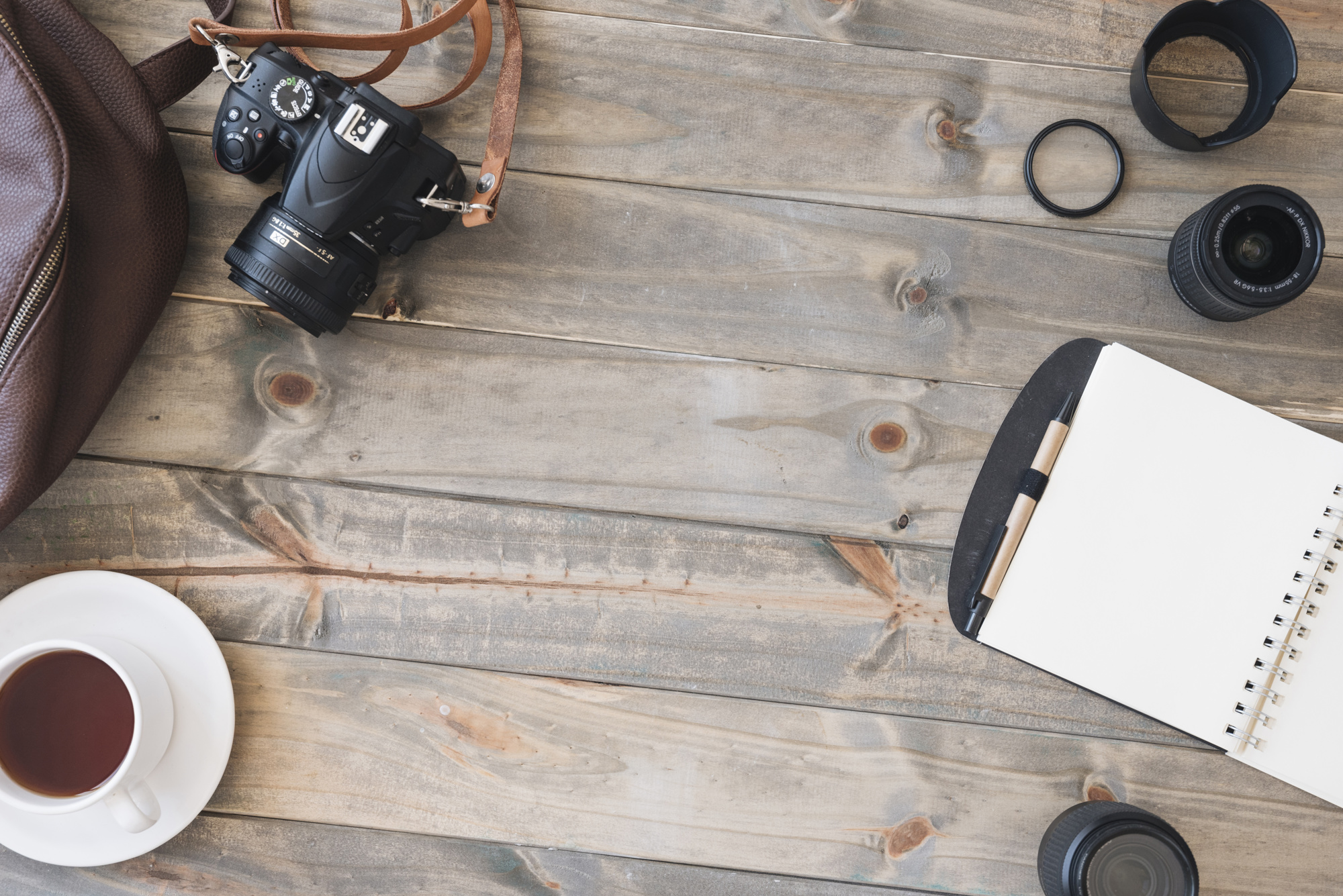 デジタル一眼レフカメラの上面図。紅茶1杯;スパイラルメモ帳。ペン;カメラのレンズと木製のテーブルの上にバッグ