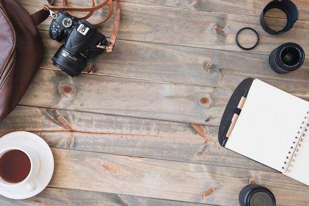 Top view of dslr camera; cup of tea; spiral notepad; pen; camera lens and bag on wooden table