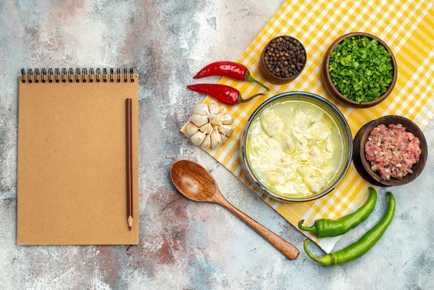 Top view dushbara dumplings soup in a bowl garlic hot peppers wooden spoon bowls with meat pepper and greens a notebook on nude table