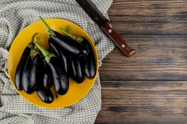 Top view of eggplants in plate with knife on cloth on left side and wooden background with copy space