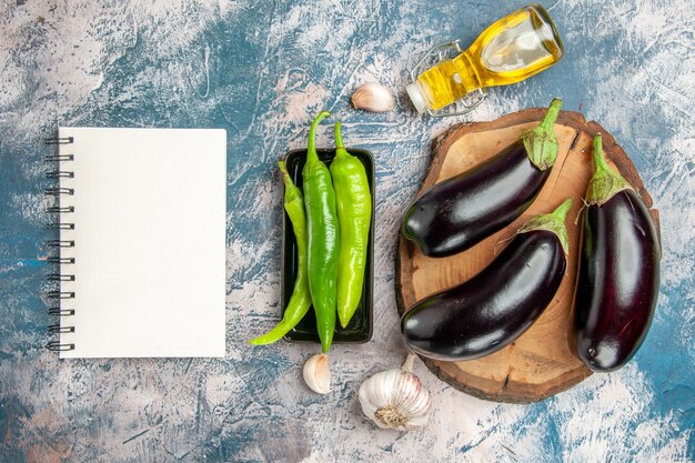 Top view eggplants on tree wood board hot peppers on black plate garlic oil a notebook on blue-white background