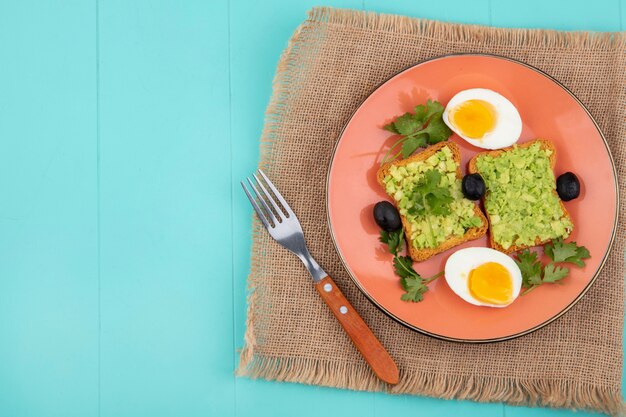 Top view of eggs with toasted slices of breads with avocado pulp onn orange plate with fork on sack cloth on blue