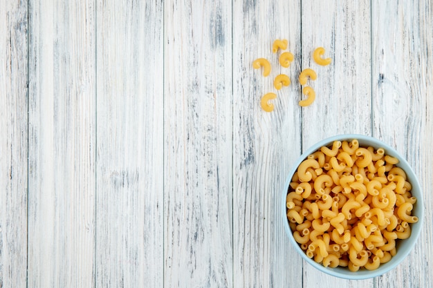 Top view elbow macaroni pasta in bowl on the right with copy space on white wooden background