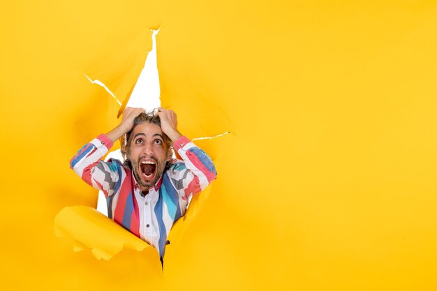 Top view of an emotional angry and exhausted young guy through a torn hole in yellow paper