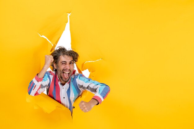 Top view of an emotional and crazy young guy posing for camera through a torn hole in yellow paper
