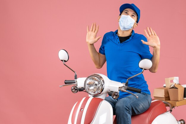 Top view of emotional delivery guy in medical mask wearing hat sitting on scooter and looking up on pastel peach background