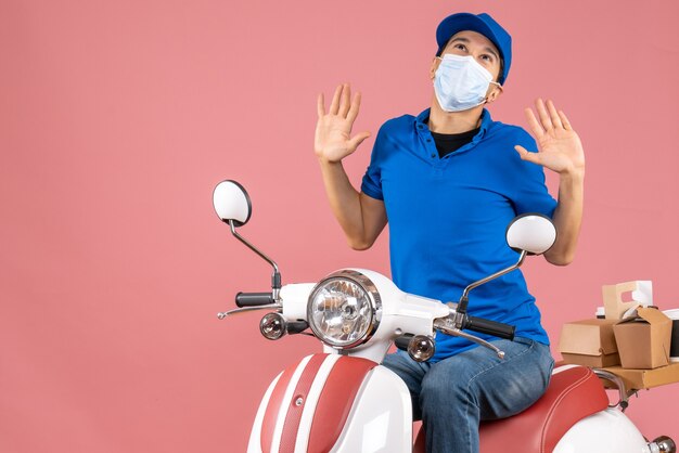 Вид сверху эмоционального доставщика в медицинской маске в шляпе, сидящего на скутере и смотрящего на пастельный персиковый фон
