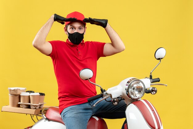 Вид сверху эмоционального молодого человека в красной блузке и шляпных перчатках в медицинской маске, доставляющего заказ, сидящего на скутере, сосредоточенного на чем-то