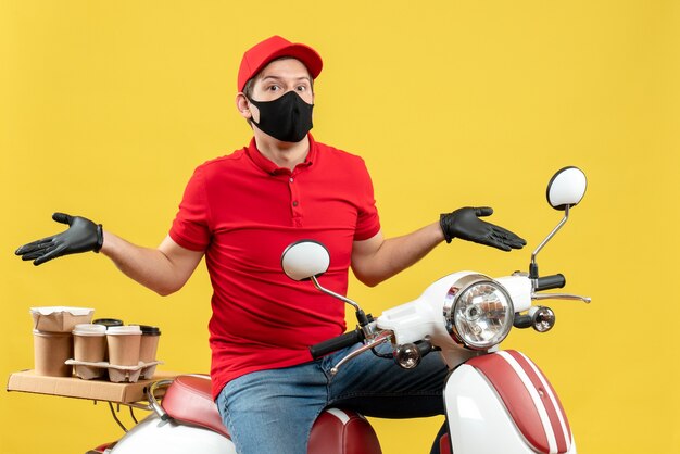 Вид сверху эмоционального молодого человека в красной блузке и шляпных перчатках в медицинской маске, доставляющего заказ, сидя на скутере, чувствуя замешательство