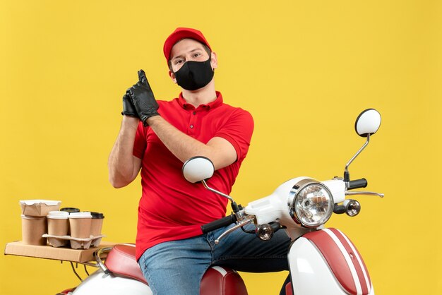 Top view of emotional young adult wearing red blouse and hat gloves in medical mask delivering order sitting on scooter making gun gesture