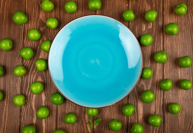 Top view of an empty blue plate and sour green plums arranged around on wooden table
