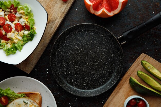 Top view of empty frying pan with vegetable salad cut grapefruit cucumber slices on black background