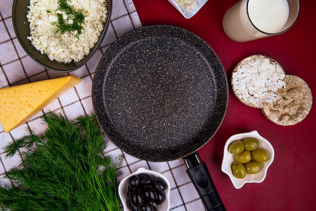 Top view of an empty pan and a piece of cheese with pickled olives and rice cakes on plaid fabric on red