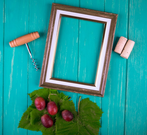 Top view of an empty picture frame with bottle screw , wine corks and sweet grapes with green grape leaves on blue wooden table