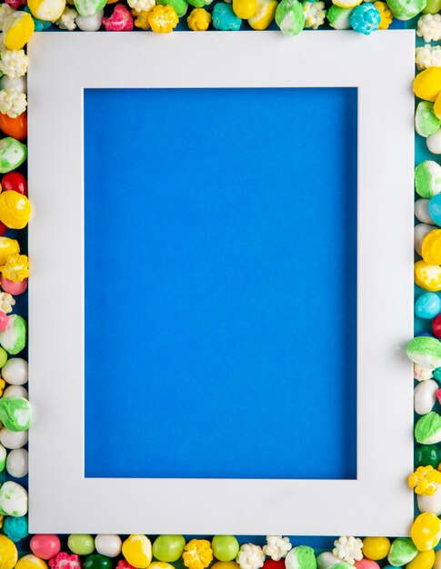 Top view of an empty picture frame with colorful candies arranged around on blue background