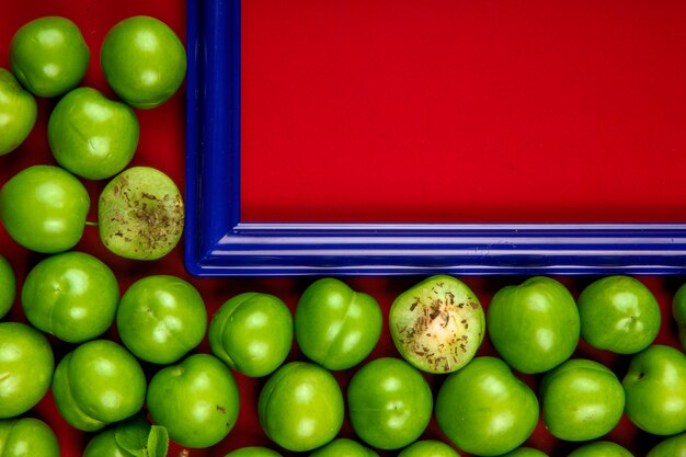 Top view of an empty picture frame with sour green plums arranged around on red table with copy space