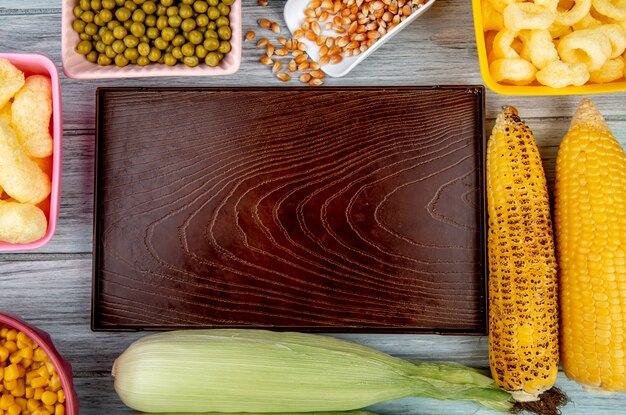 Top view of empty tray with green peas corn seeds corn pop cereals and corn cobs on wooden surface