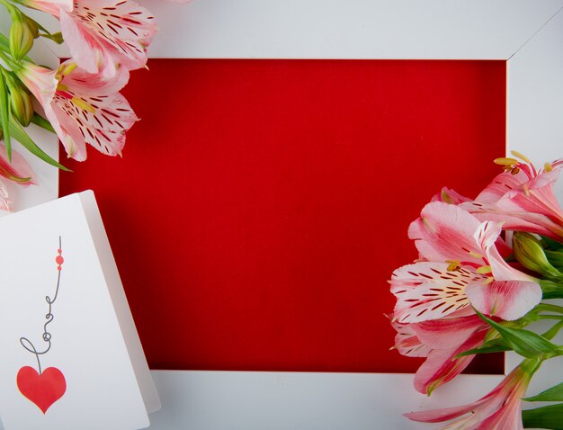 Top view of an empty white picture frame with pink color alstroemeria flowers and a postcard on red background with copy space