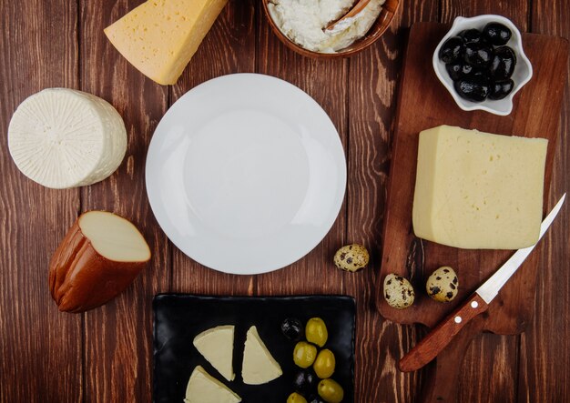 Top view of an empty white plate and different kind of cheese with pickled olives and quail eggs arranged on rustic table