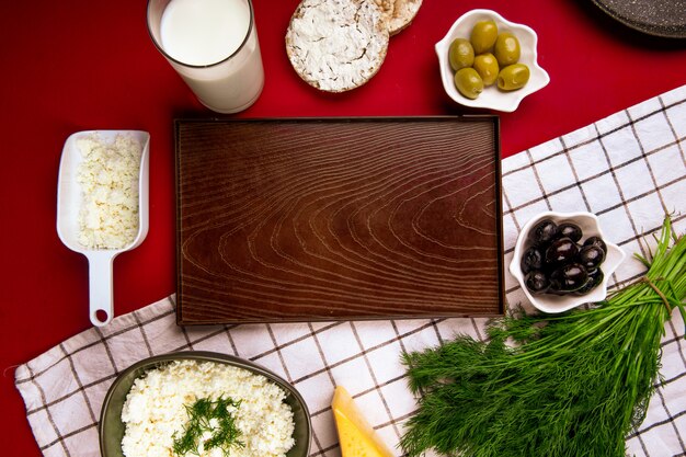 Top view of an empty wood tray and cottage cheese in a bowl with pickled olives dill and rice cakes on plaid fabric on red