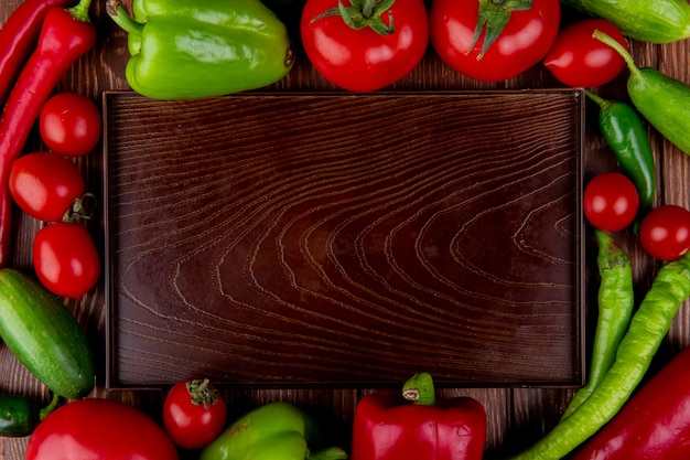 空の木製トレイと新鮮な野菜の完熟トマトの緑と赤の唐辛子とカラフルなピーマンの素朴な平面図