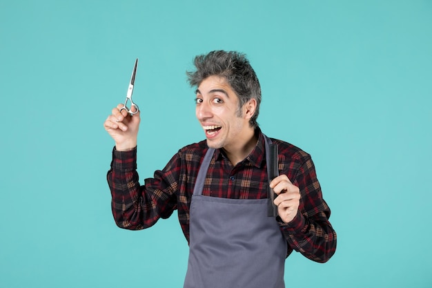 Top view of excited young hairdesser wearing gray apron and holding comb and scissor on blue color background