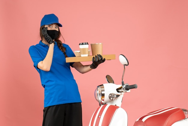 Top view of fearful courier girl wearing medical mask gloves standing next to motorcycle holding coffee small cakes on pastel peach color background