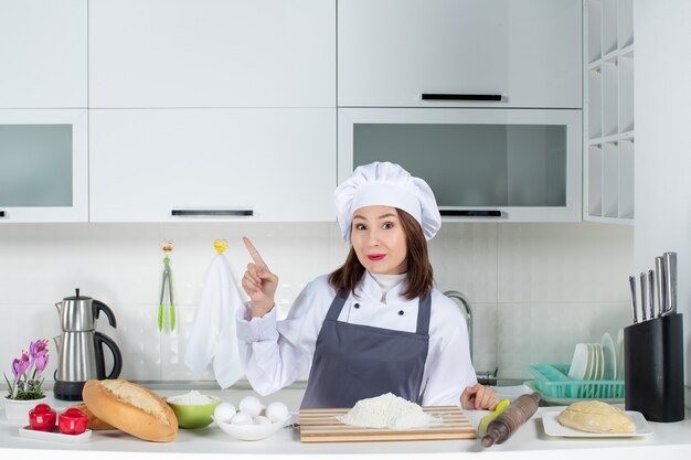 Top view of female chef in uniform standing behind the table with cutting board bread vegetables pointing up in the white kitchen