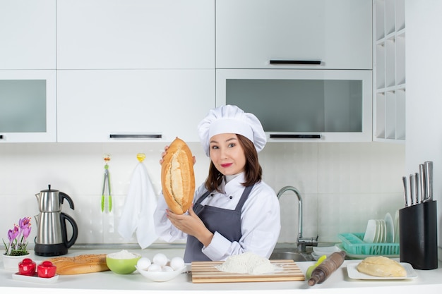 Top view of female chef in uniform standing behind the table with cutting board and foods showing bread in the white kitchen