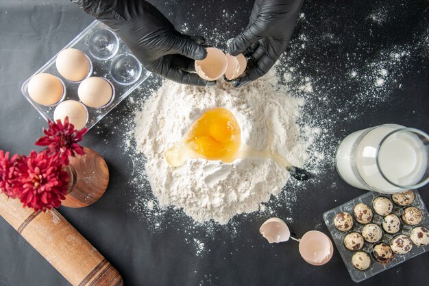 Top view female cook breaking eggs into flour on dark surface