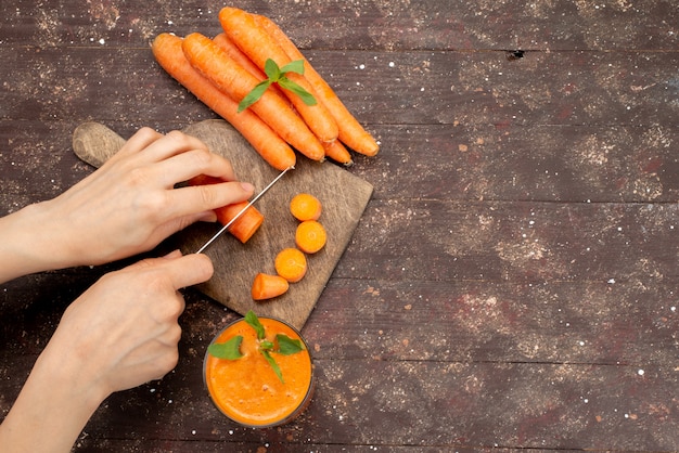 Бесплатное фото Вид сверху женщина резки морковь на коричневый деревянный стол вместе со свежим морковным соком на коричневый