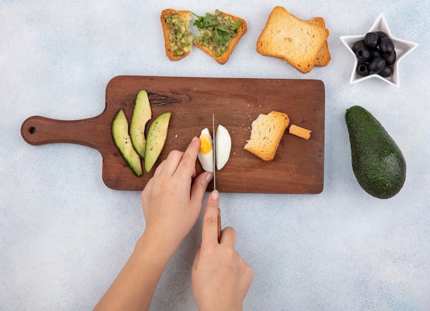 Free photo top view of female hand cutting boiled egg into slices on wooden kitchen board with avocado slices toasted slice of bread black olives on white