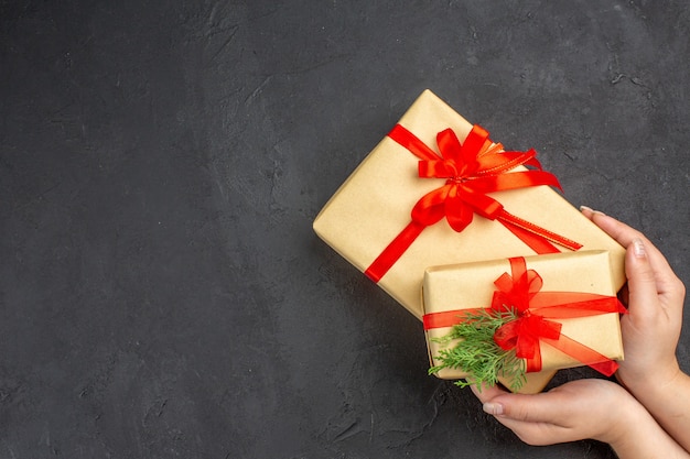 Top view female hand holding big and small xmas gifts in brown paper tied with red ribbon fir branch on dark background with free space